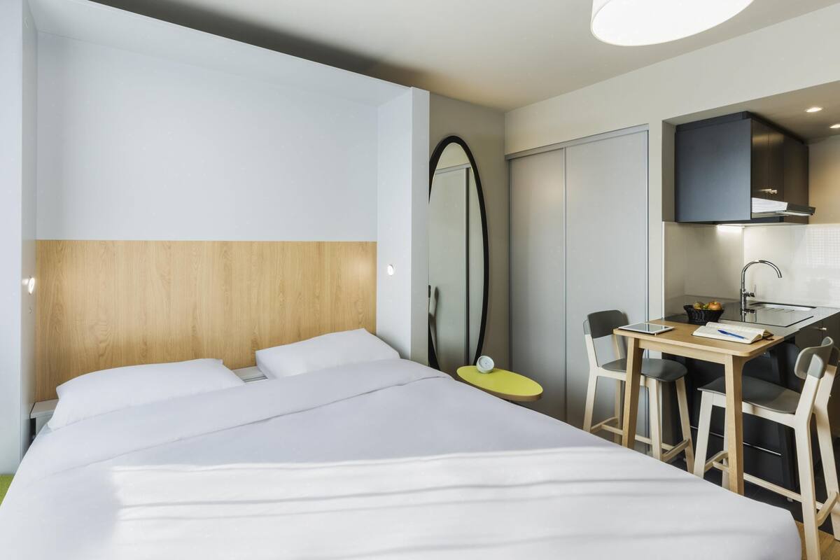 A hotel room with a bed, a desk with a chair, a mirror, and a kitchenette.