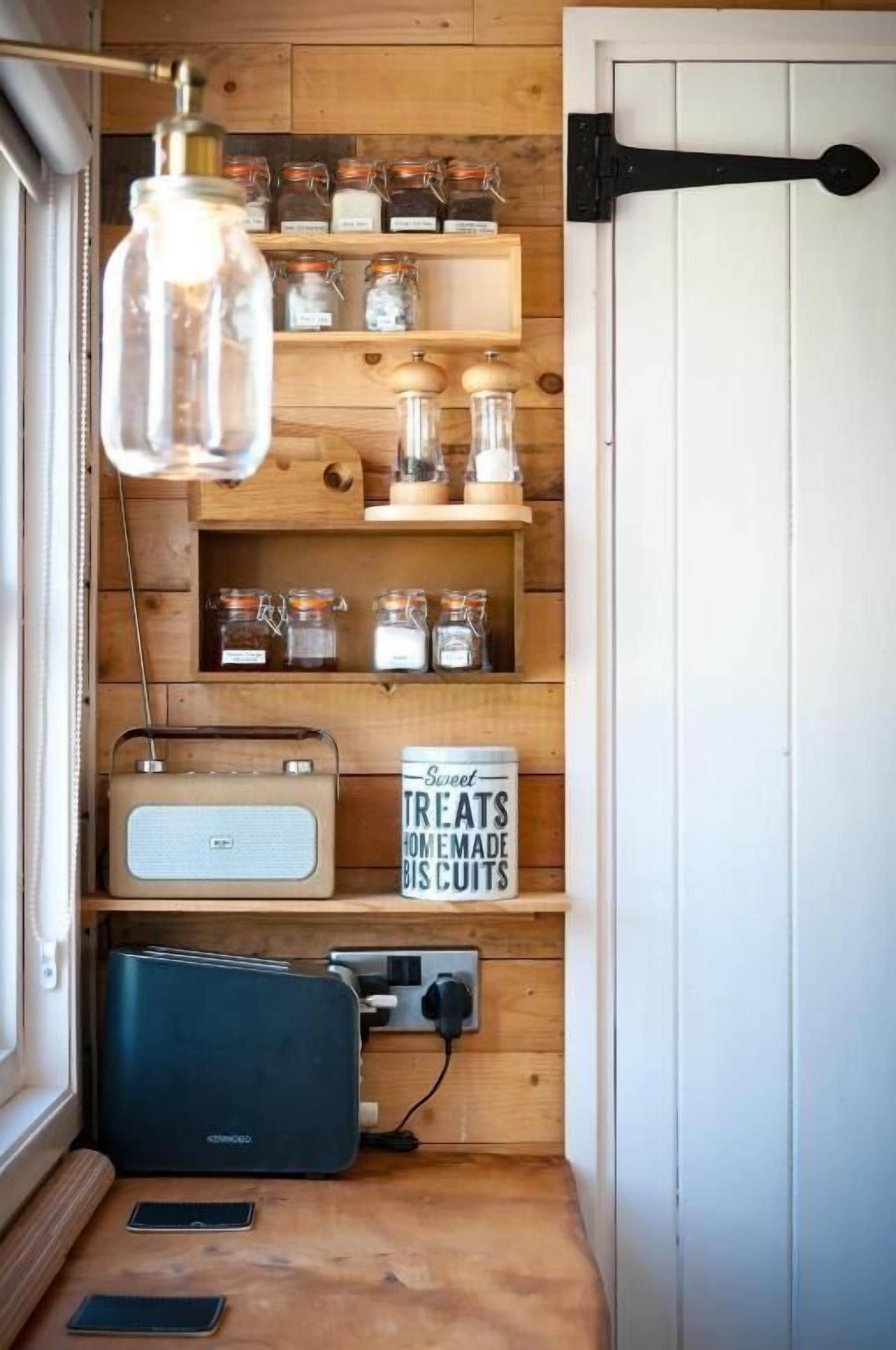 A compact kitchen with wooden shelves, jars, a vintage radio, and a sign about homemade treats.