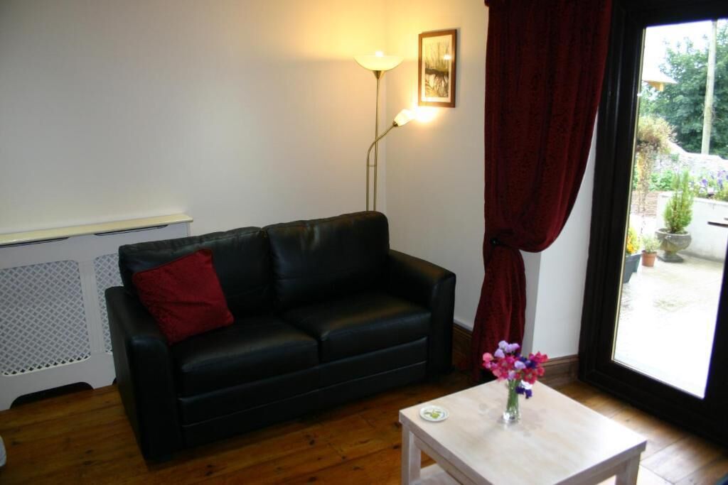 A black leather sofa with a red pillow, a small white table with a vase of flowers, a floor lamp, and a window with red curtains.