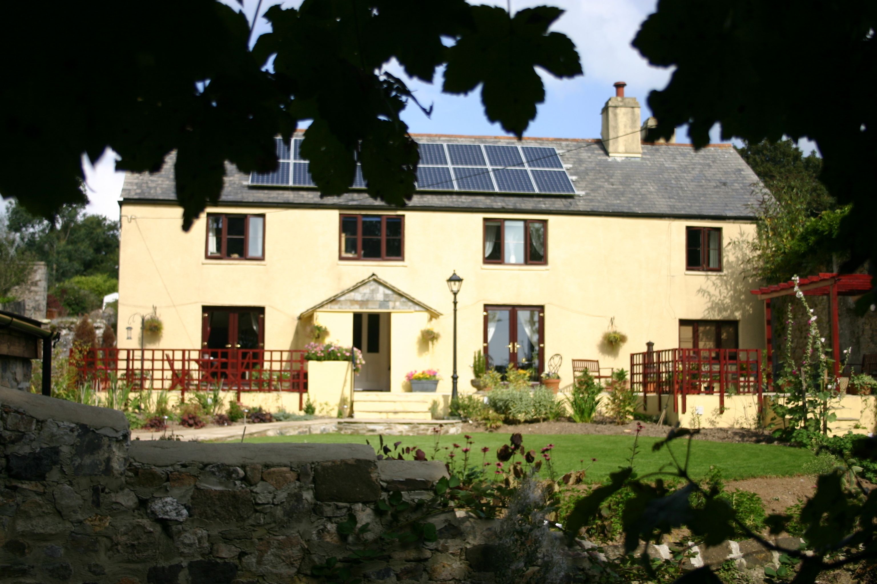 A two-story house with a stone wall in the foreground, a small garden with various plants, and a solar panel on the roof.