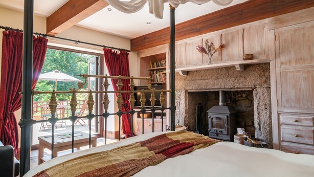 A bedroom with a large bed, a stone fireplace, wooden beams, and a view of the outdoors. - Cadleigh Manor B&B (Ivybridge)