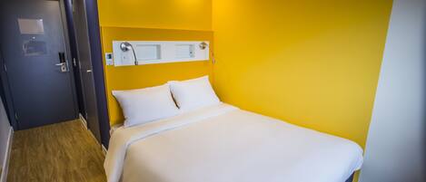 Una habitación de hotel con una cama, suelo de madera, una puerta y una pared con interruptor de luz.