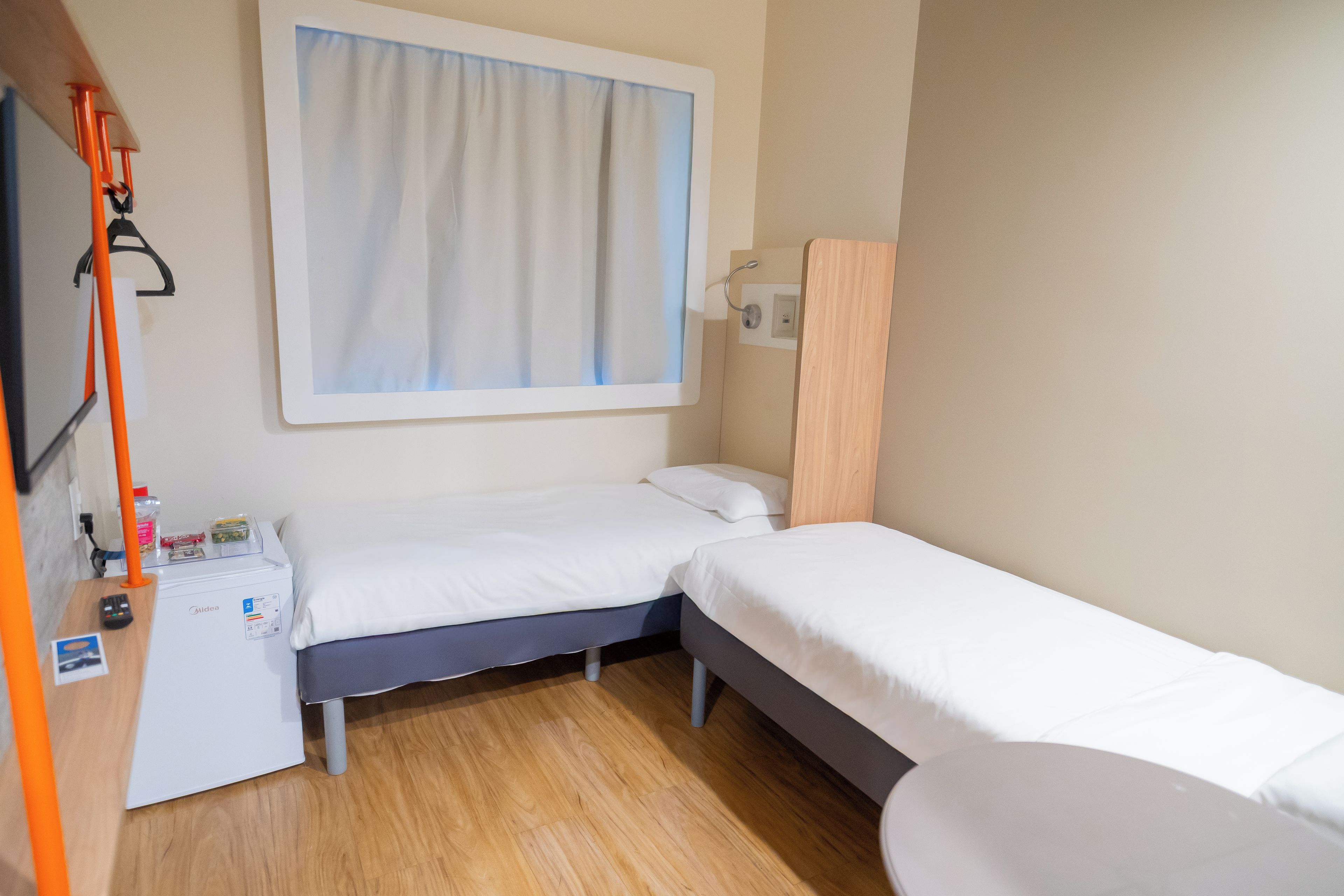 A small, compact room with a single bed, a desk with a computer, and a window with curtains.