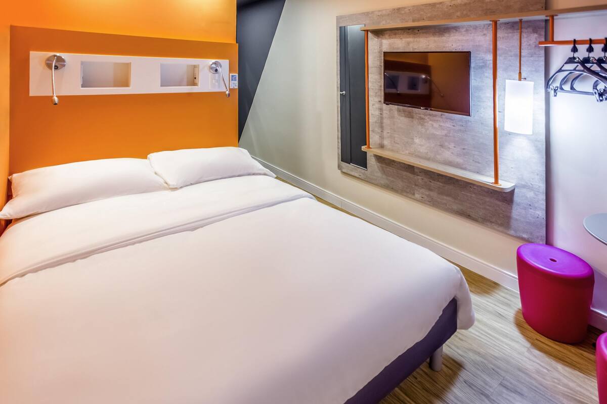 A modern hotel room with a large bed, a wooden accent wall, a small table, and a red stool.