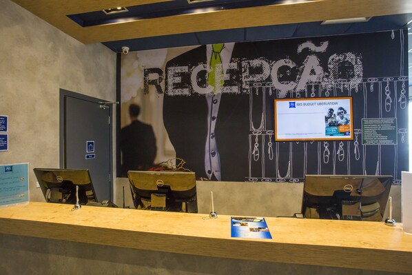 A reception desk with a wooden counter, two chairs, and a Dell computer. A poster with the word "RECLIPCAO" is on the wall.