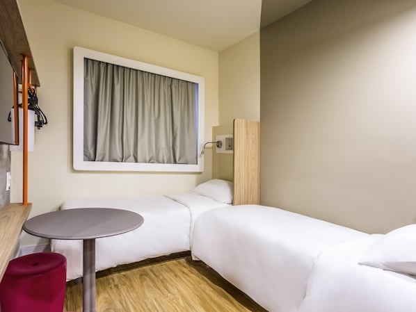 Quarto de hotel com uma cama, uma pequena mesa redonda, um banquinho vermelho e uma janela com cortinas.