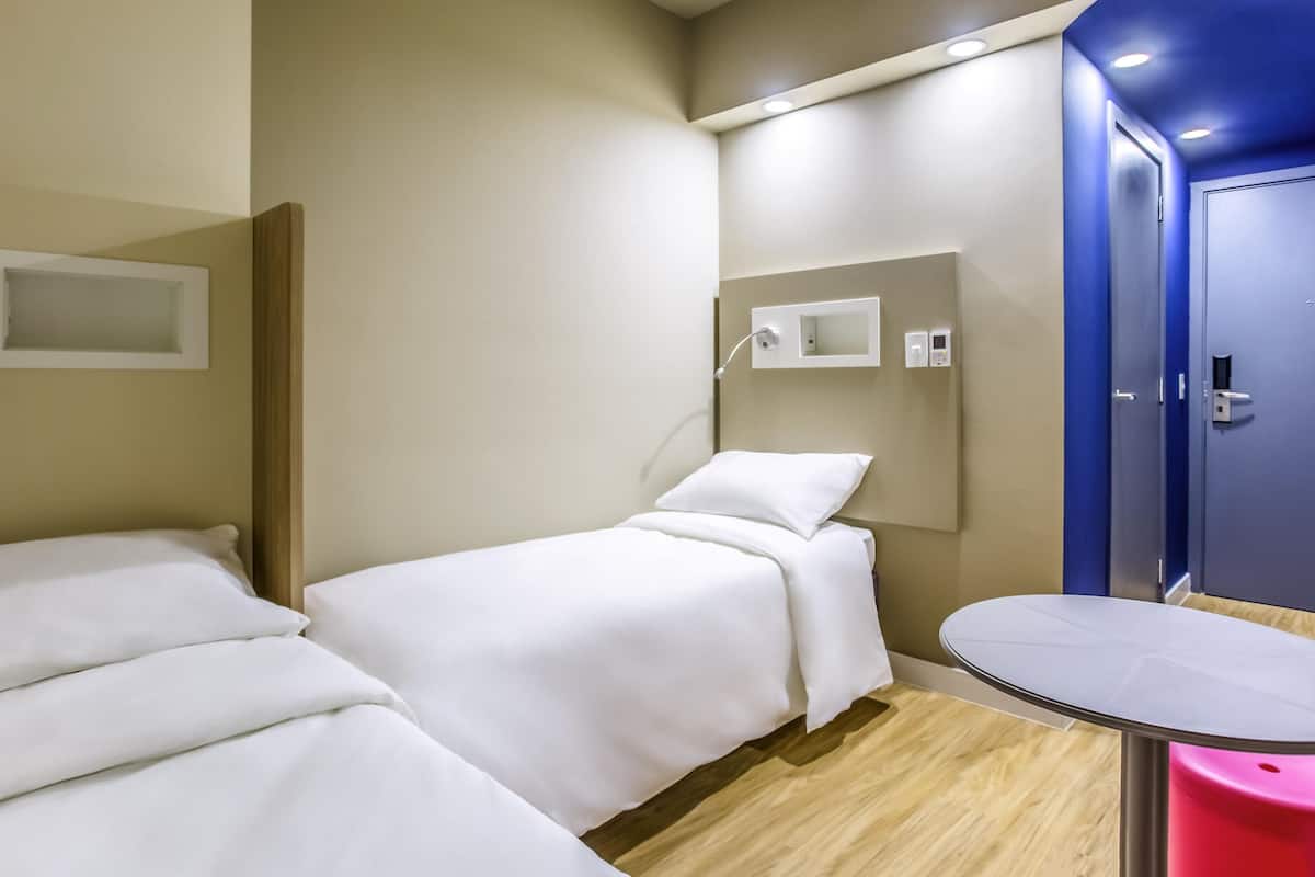 A hotel room with two beds, a small table, a mirror, and a blue door.