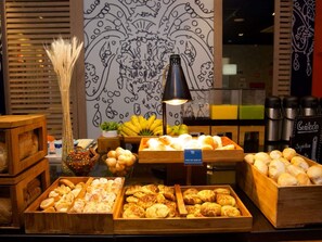 Un buffet de petit-déjeuner proposant diverses pâtisseries et des fruits, un mur décoratif orné de motifs abstraits et une lampe noire.