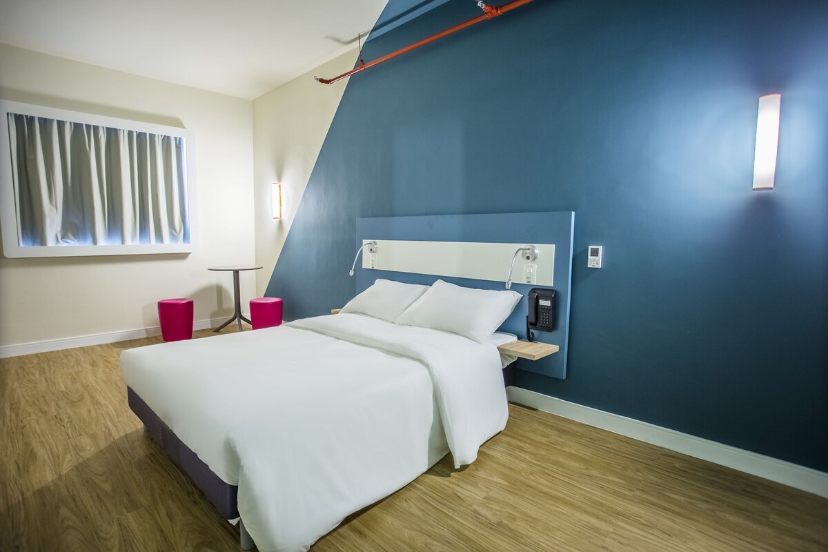 A modern hotel room with a bed, a bedside table, a lamp, and a window with curtains.