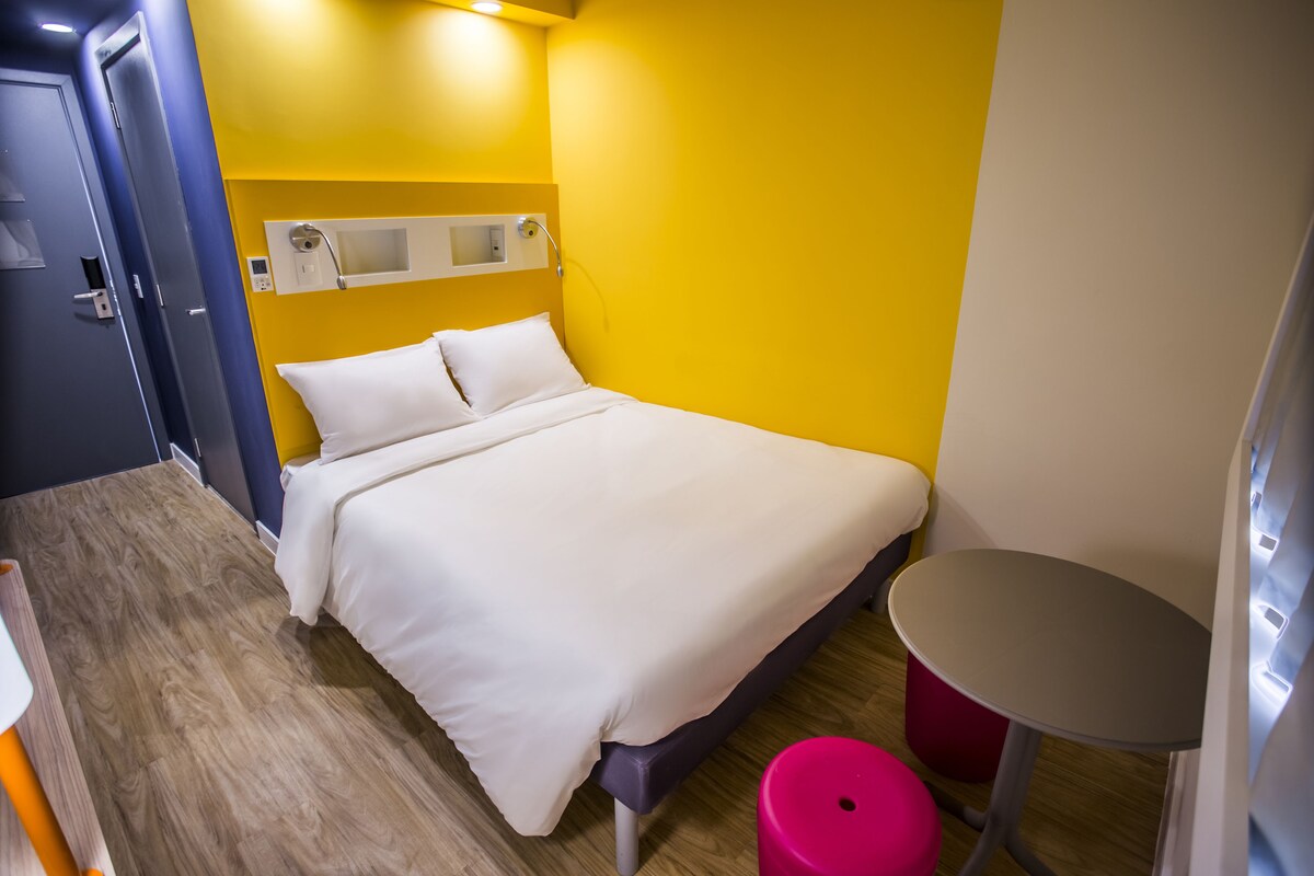 A room with a bed, a small table, and a red stool.