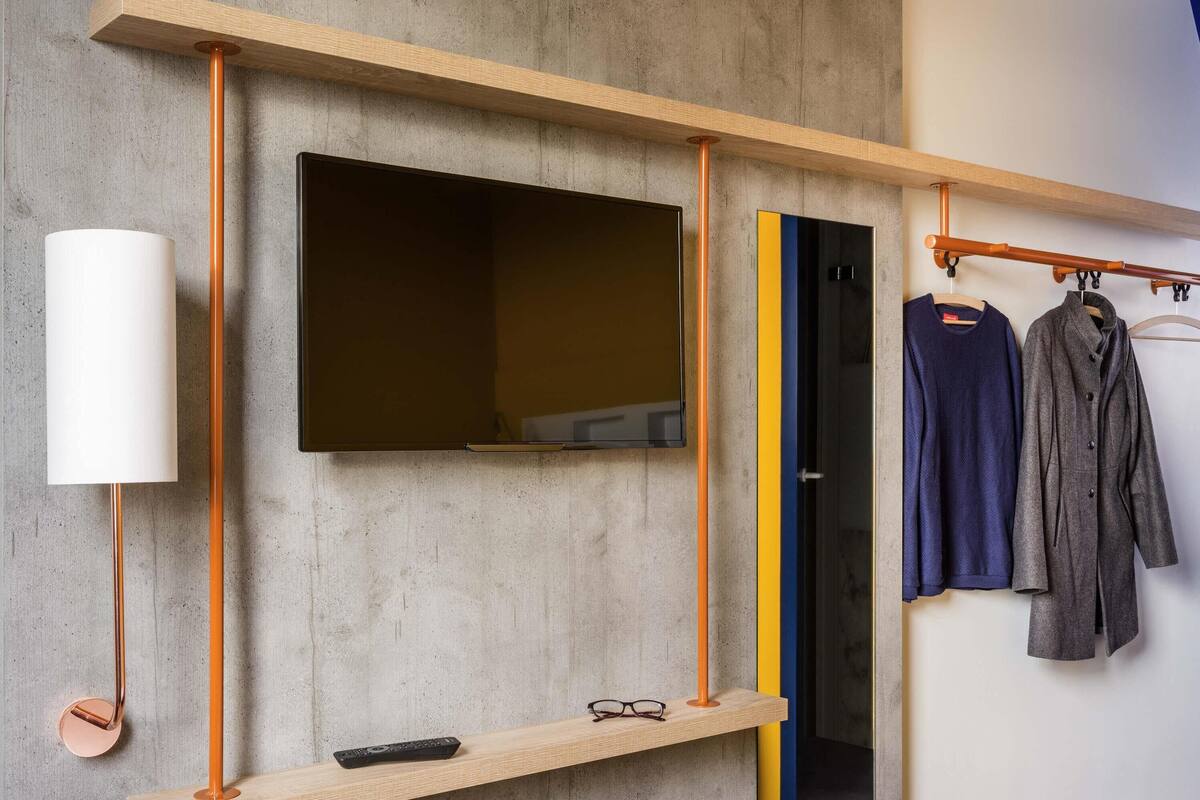 Una habitación moderna con un televisor montado, una estantería de madera con un control remoto y un perchero con dos prendas.