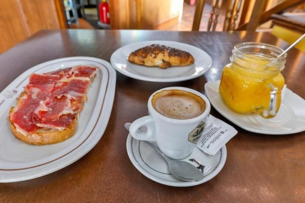 Una mesa con jamón serrano, un croissant, una taza de café y un tarro de jugo de naranja.