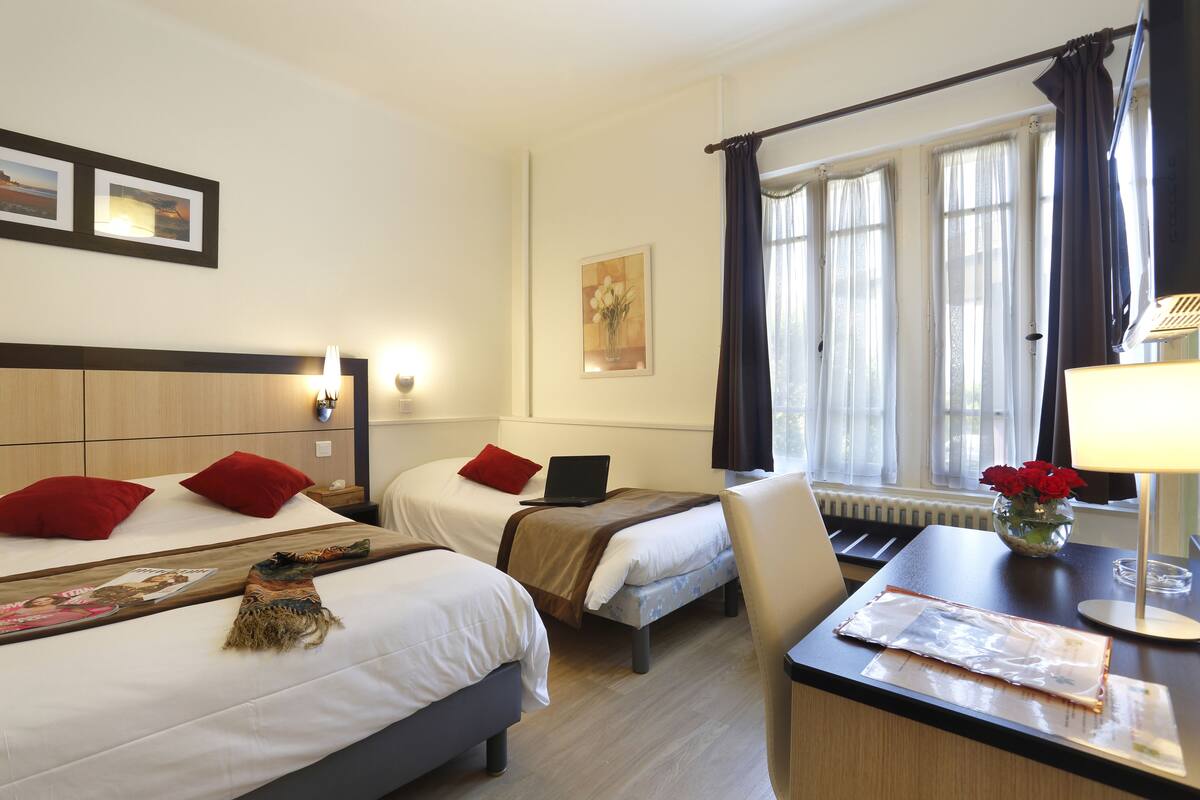 A hotel room with two beds, a desk, a lamp, a vase with flowers, and a window with curtains.