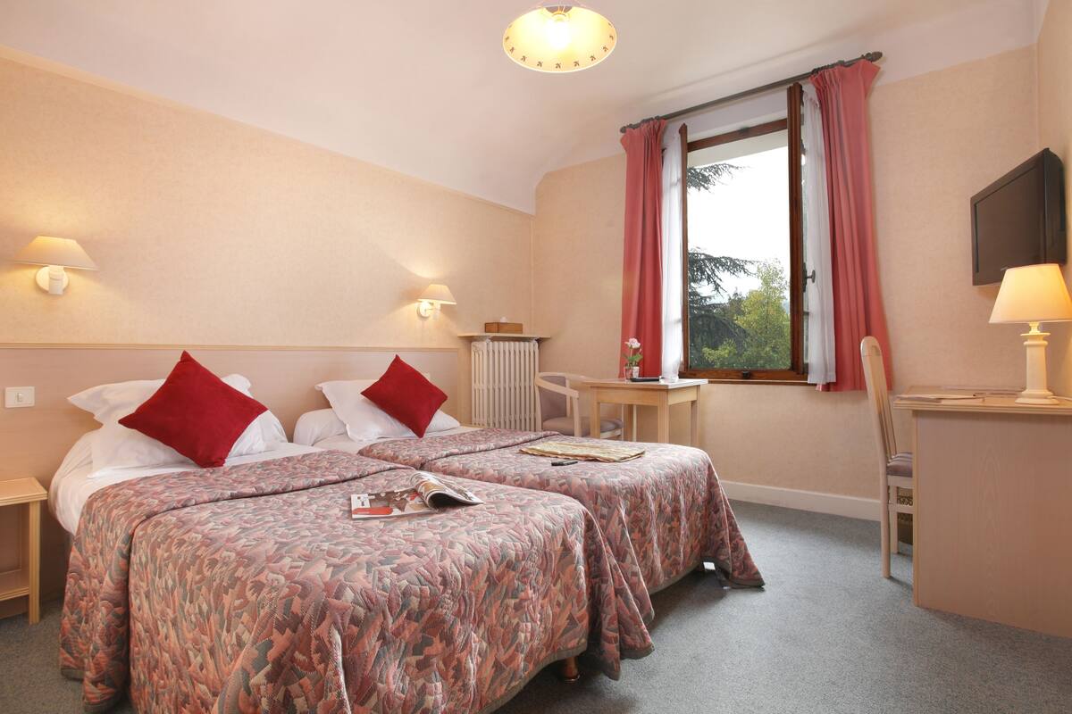 A hotel room with two beds, a desk, a chair, and a window with curtains.
