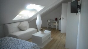 Un dormitorio pequeño estilo loft, de color blanco, con techo inclinado, un sofá blanco, una mesita pequeña y un televisor montado en la pared.