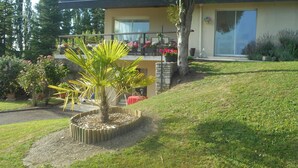 Una casa de dos pisos con un balcón, un jardín con una palmera y diversas flores.