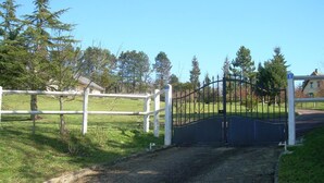 Una entrada cerrada con un portón metálico, una valla blanca y un camino de grava que conduce a una zona verde con árboles y una casa al fondo.