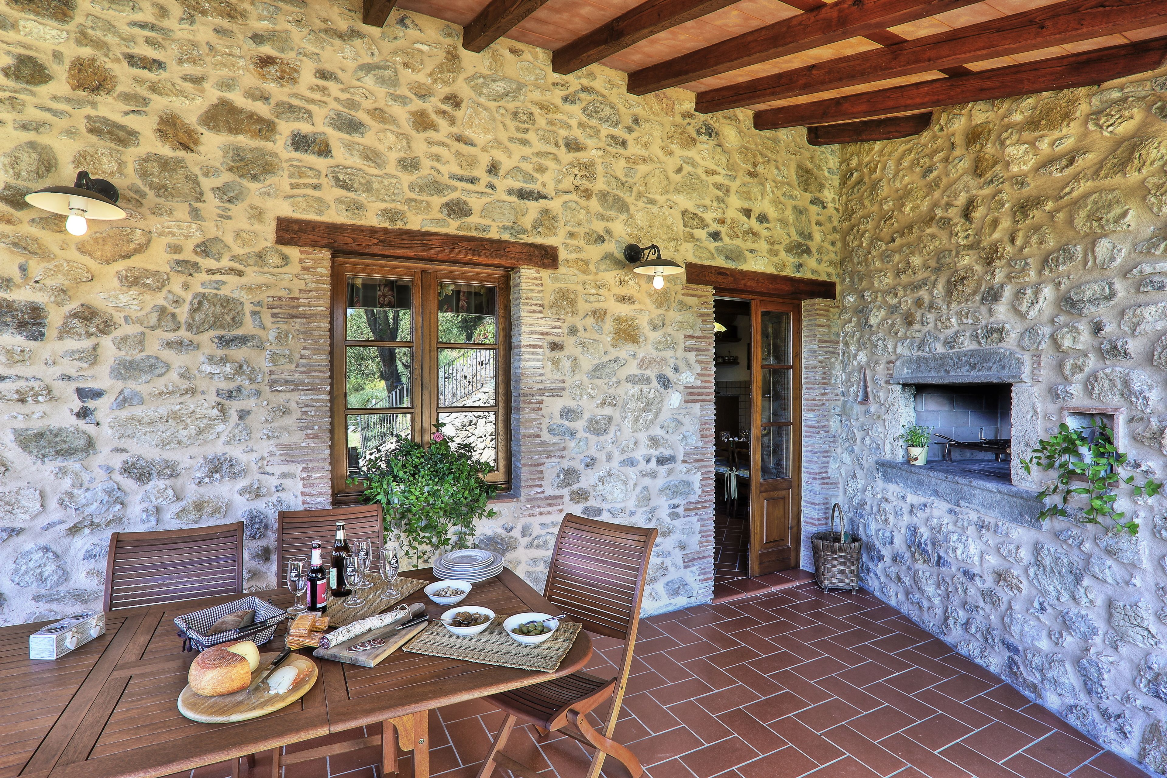 A stone-walled patio with a wooden table set for dining, a stone fireplace, and wooden beams on the ceiling.