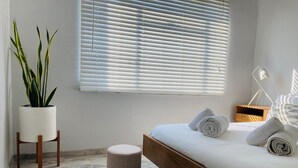 A modern bedroom with a wooden bed frame, a small round ottoman, a potted plant, and a bedside table with a lamp.