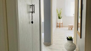 A hallway with a light-colored wooden door, a white wall, and a grey wall. There is a potted plant and a small stool in the distance.