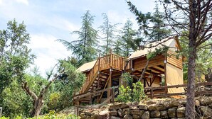 Un chalet rustique avec un escalier en bois menant à une terrasse, entouré d’arbres et d’un mur de pierres.