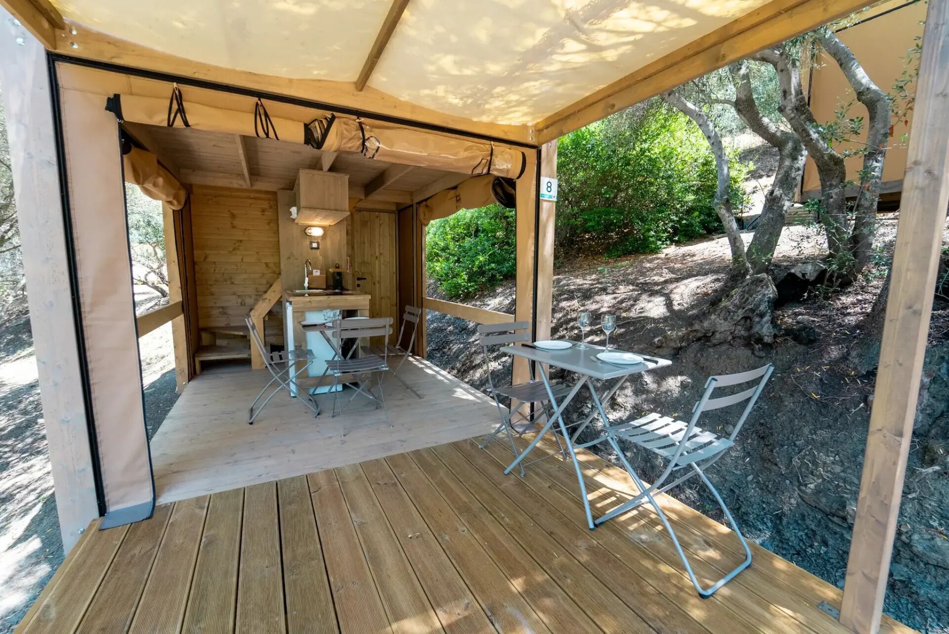 Una terraza de madera con mesa y sillas, una kitchenette pequeña, y vistas a un paisaje rocoso con árboles.
