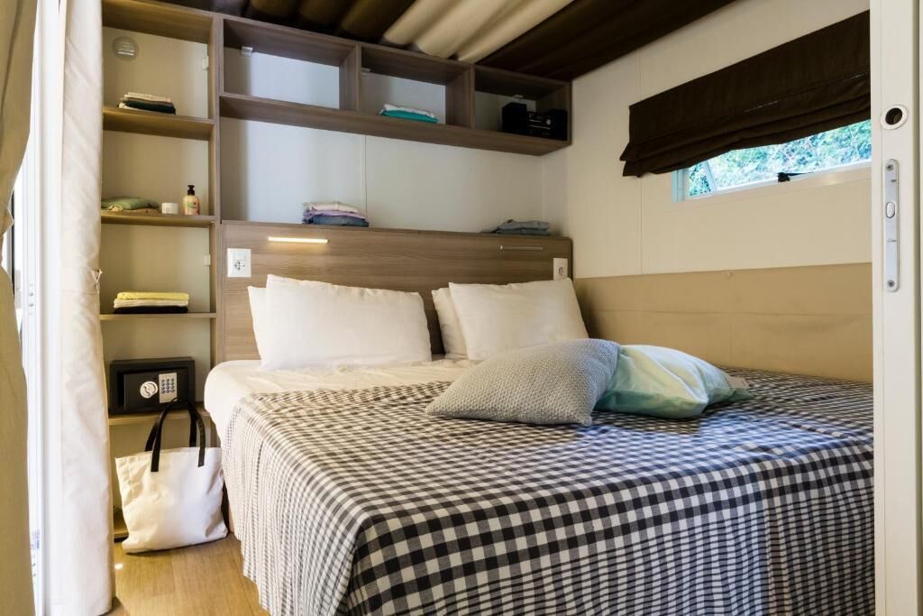 A neatly made bed with a checkered bedspread, a bedside table with a lamp, and built-in shelving with folded clothes and a bag.
