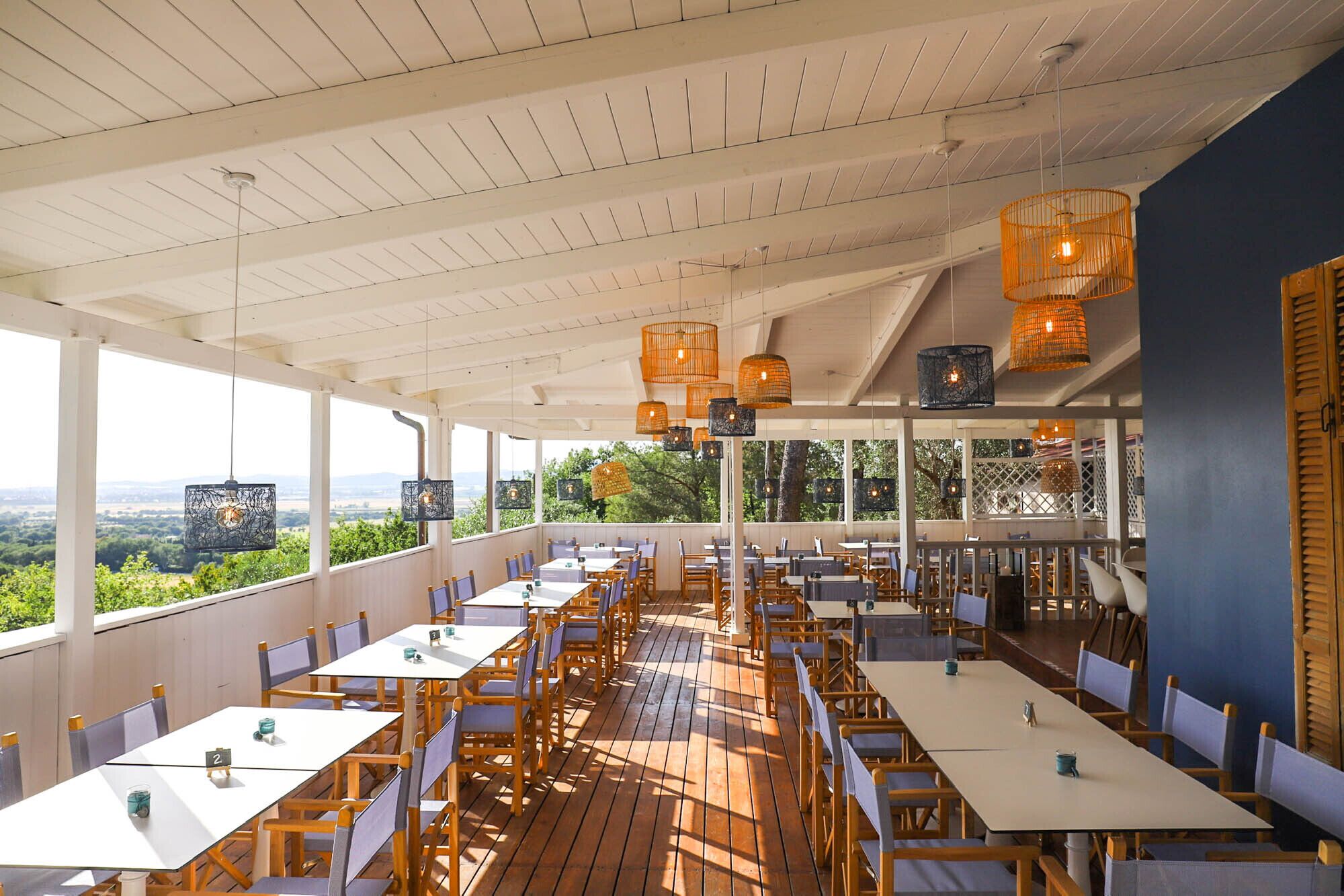An outdoor restaurant with wooden tables and chairs, hanging orange lanterns, and a view of greenery.