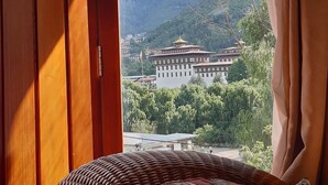 A room with a wicker bed, a view of a mountain and a building through an open door. - Kisa Villa (Thimphu)