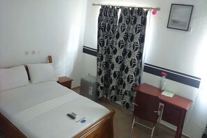 A hotel room with a bed, a desk, a chair, a small trash bin, and a curtain with a leaf pattern.