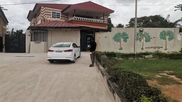 Una casa de dos pisos con techo rojo, un coche estacionado adelante y un muro con murales de palmeras.