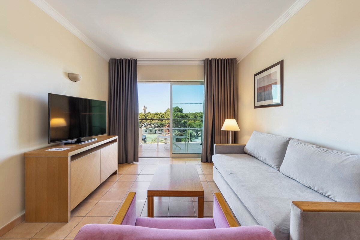 A modern living room with a flat-screen TV, a sofa, a coffee table, and a balcony with curtains.