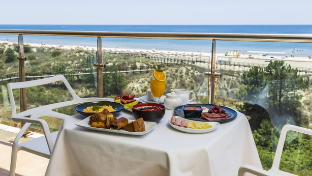 A breakfast table with various dishes and a glass of orange juice, overlooking a beach and greenery. - Hotel Apartamentos Dunamar (Vila Real Santo Antonio)