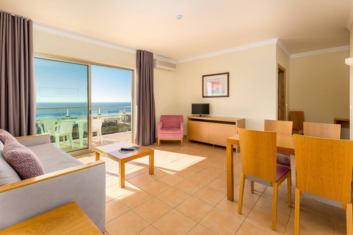 A modern hotel room with a balcony, a sofa, a dining table, and a TV.