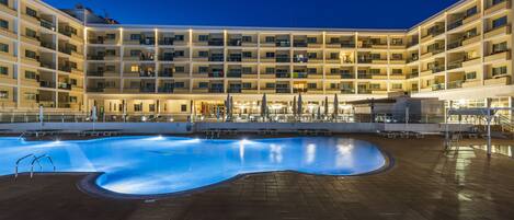 A large indoor swimming pool with a curved design, surrounded by a multi-story hotel building with balconies and a tiled floor.
