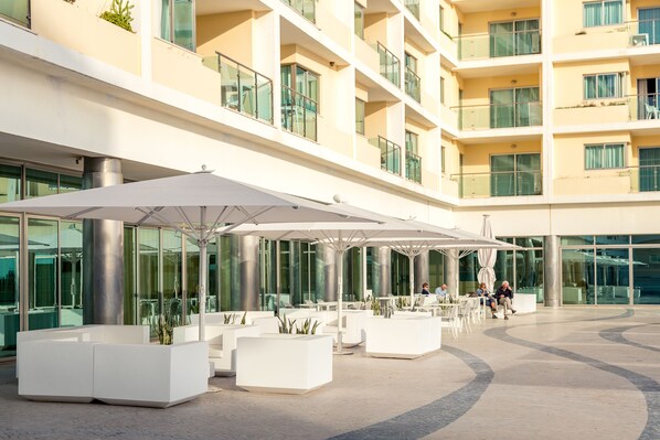 A modern outdoor seating area with white planters and umbrellas, surrounded by a multi-story building with balconies and large windows.