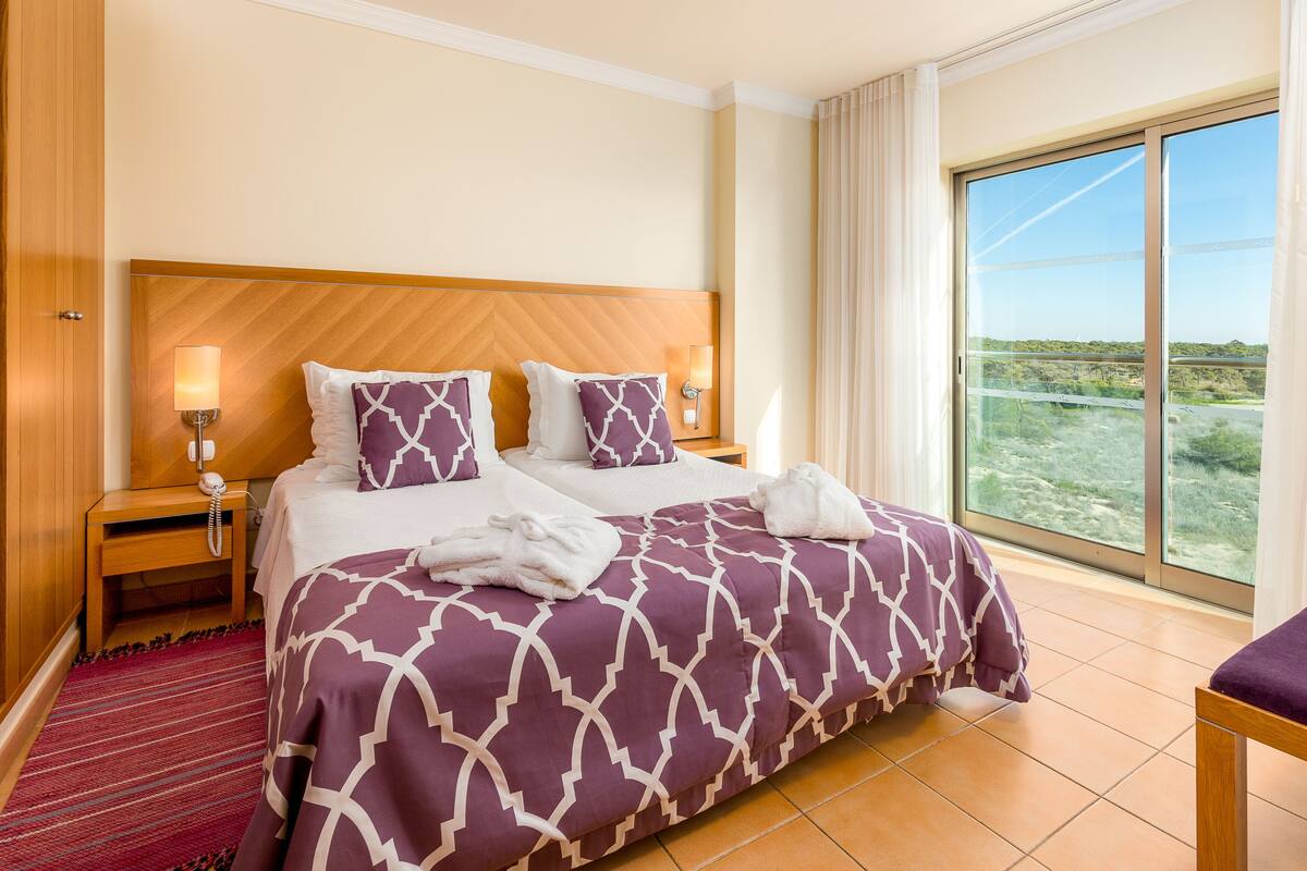 A hotel room with a large bed, two bedside lamps, a wooden headboard, a large window with a view, and a red carpet.