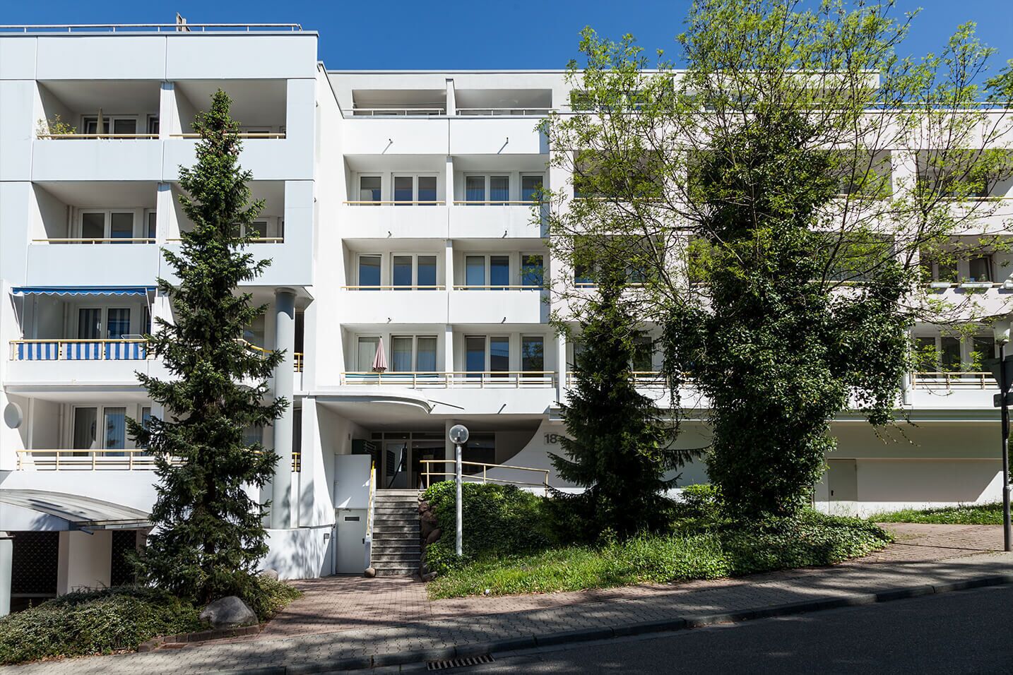 A modern multi-story building with balconies, a central entrance with steps, and surrounding greenery.