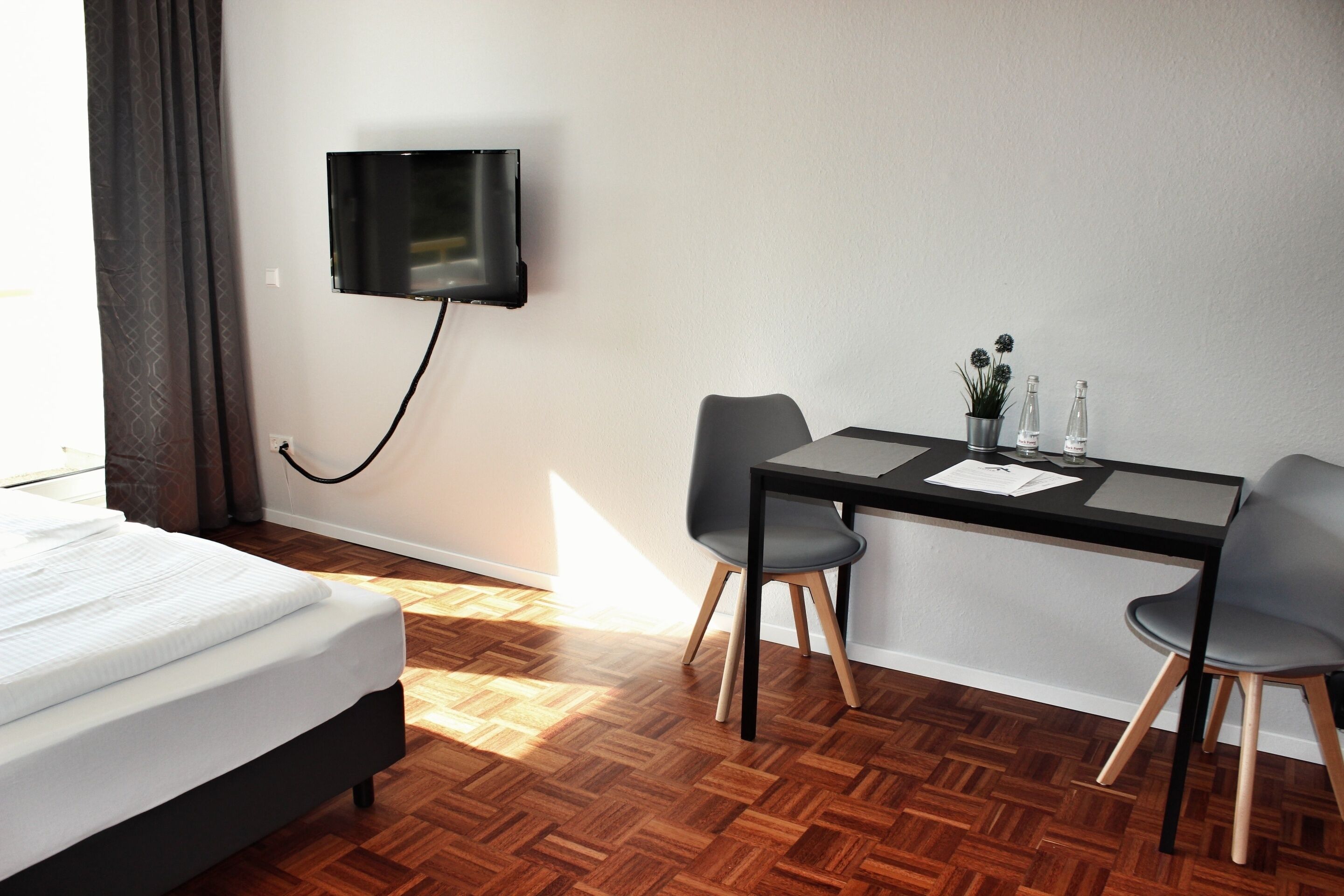 Habitación de hotel con cama, escritorio con dos sillas, televisor montado en la pared y una planta en maceta sobre el escritorio.