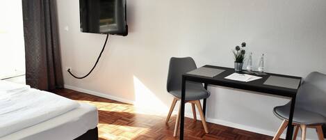 Habitación de hotel con cama, escritorio con dos sillas, televisor montado en la pared y una planta en maceta sobre el escritorio.