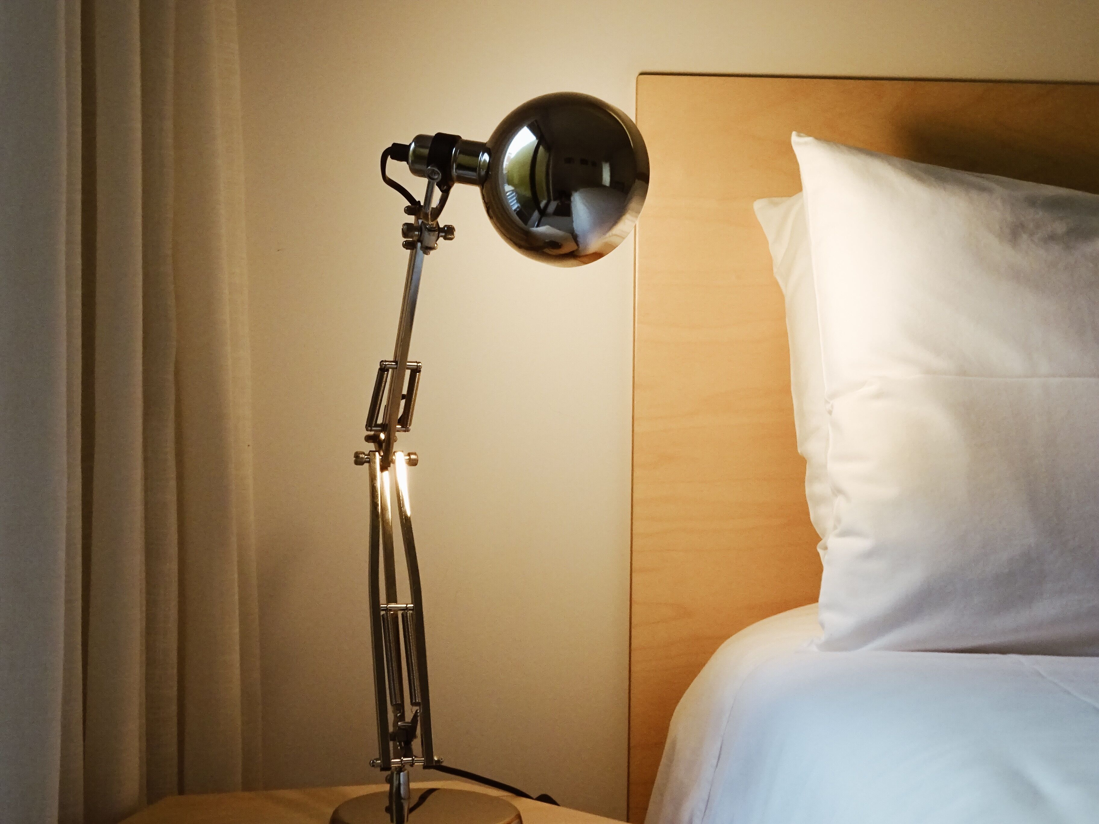 A bed with white bedding and a wooden headboard, a floor lamp with a metallic finish, and a curtain partially drawn to the side.