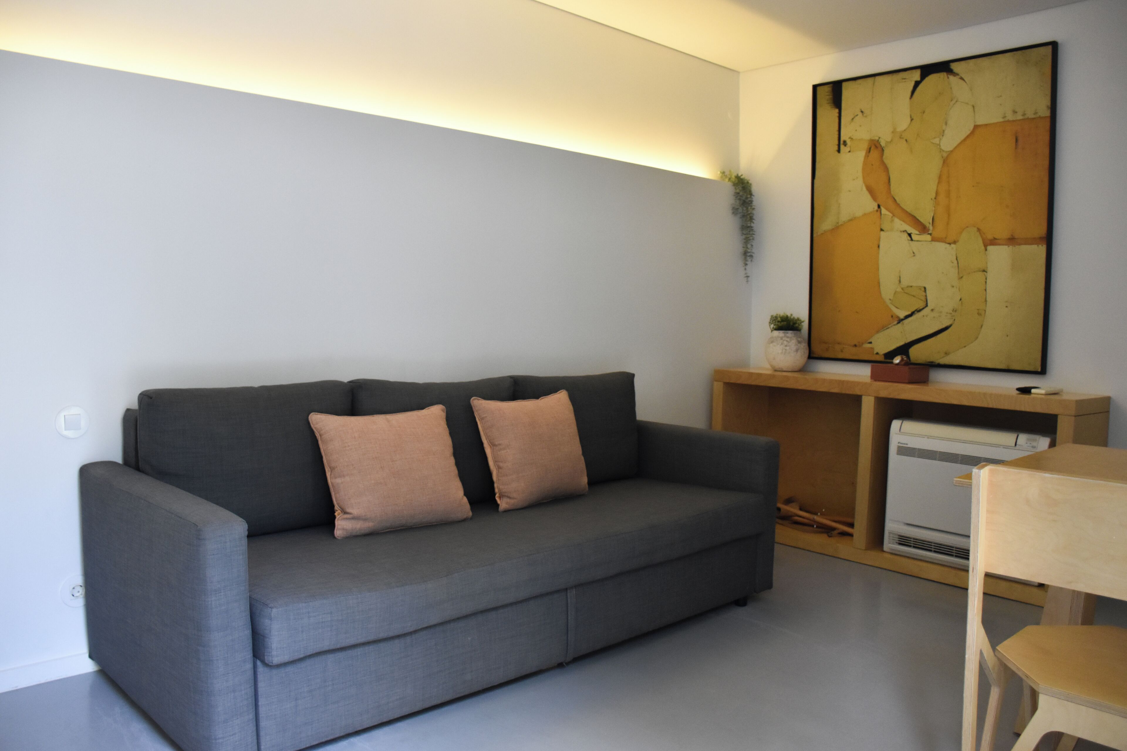 Ein modernes Wohnzimmer mit einem grauen Sofa, zwei Kissen, einem Holztischchen, einem großen abstrakten Gemälde und einer kleinen Topfpflanze.