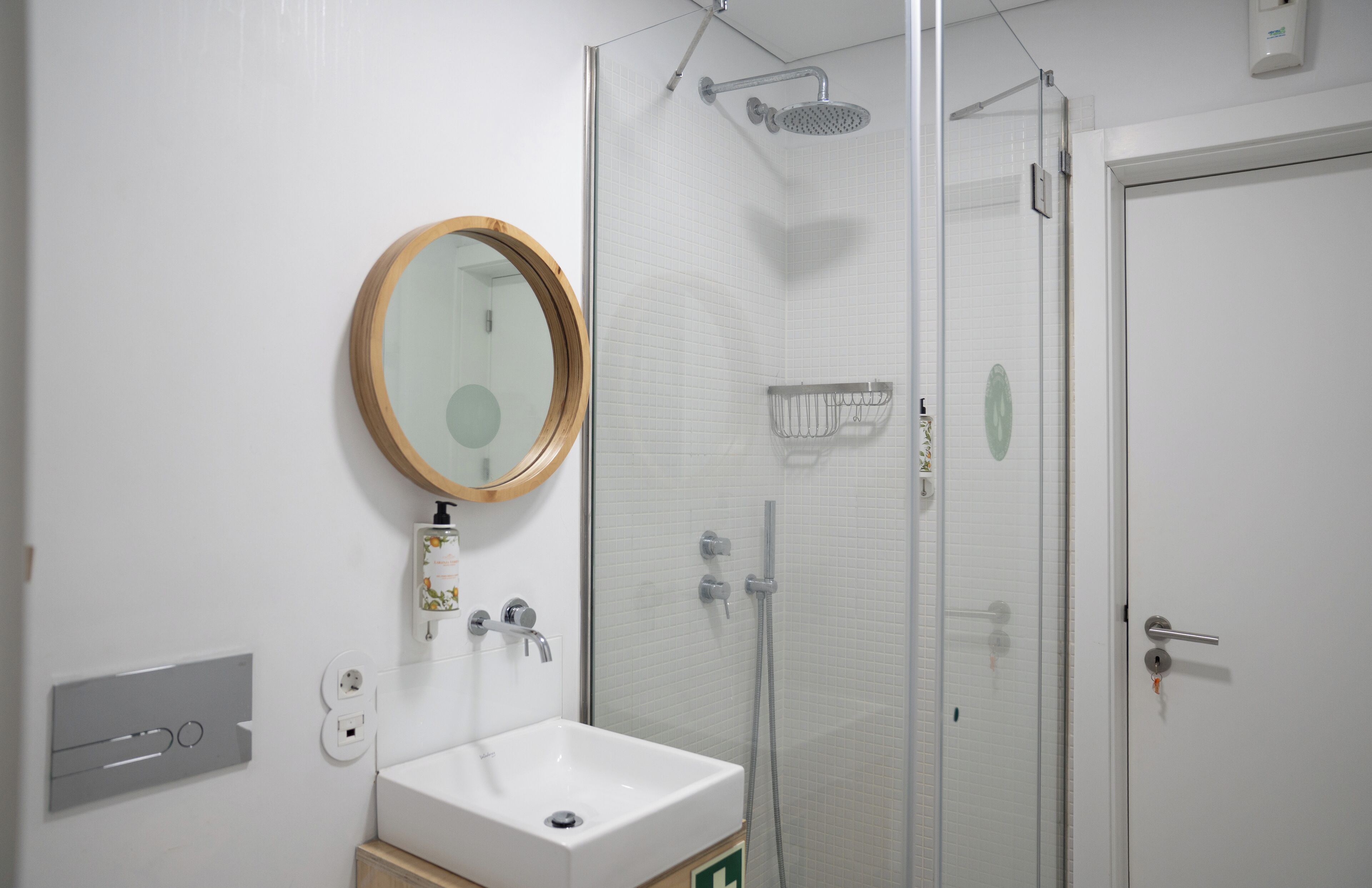 Une salle de bain avec un lavabo blanc, un miroir rond, un espace douche et une porte.