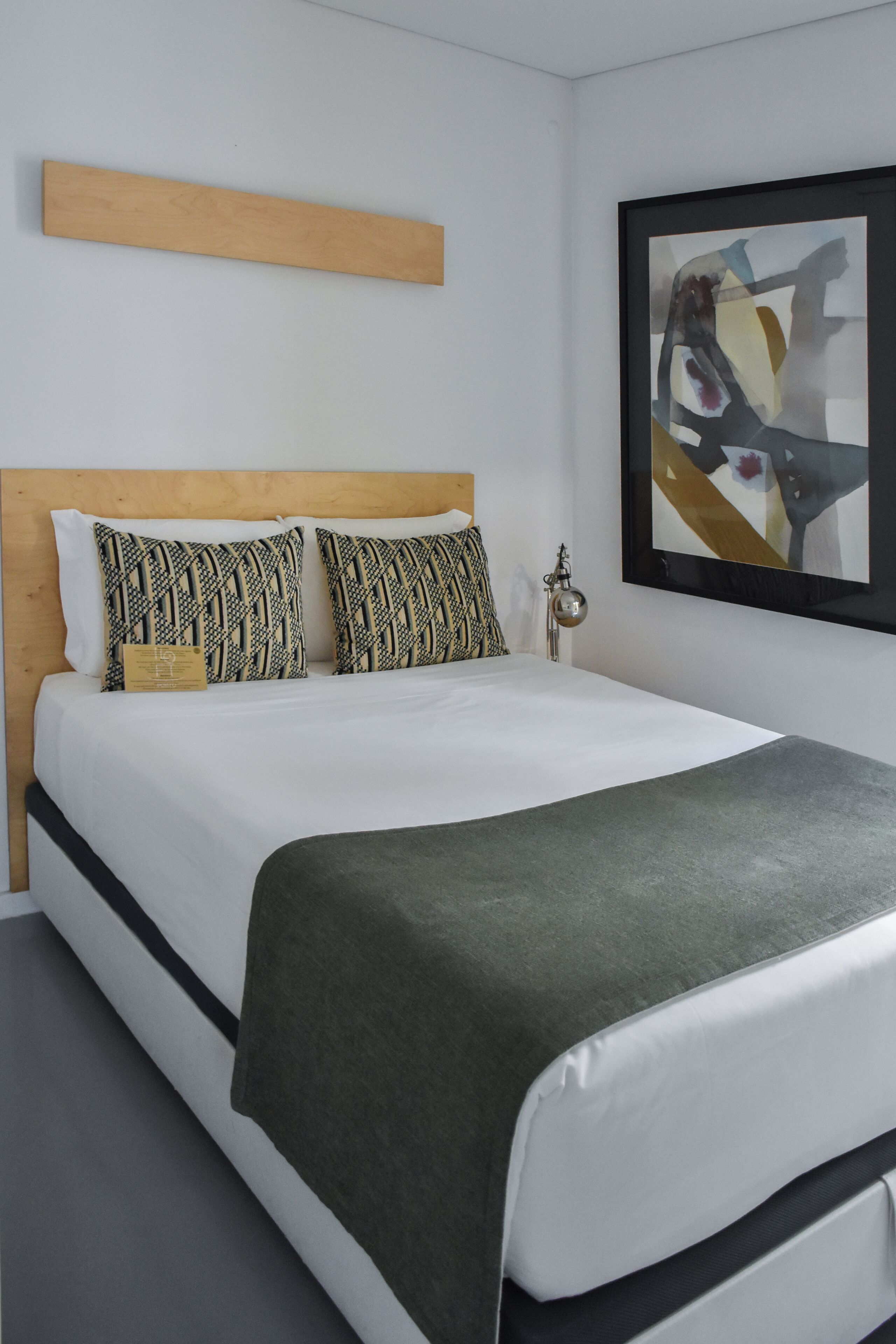 A neatly made bed with a wooden headboard, two patterned pillows, a green throw blanket, and a framed abstract painting on the wall.