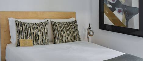 A neatly made bed with a wooden headboard, two patterned pillows, a green throw blanket, and a framed abstract painting on the wall.