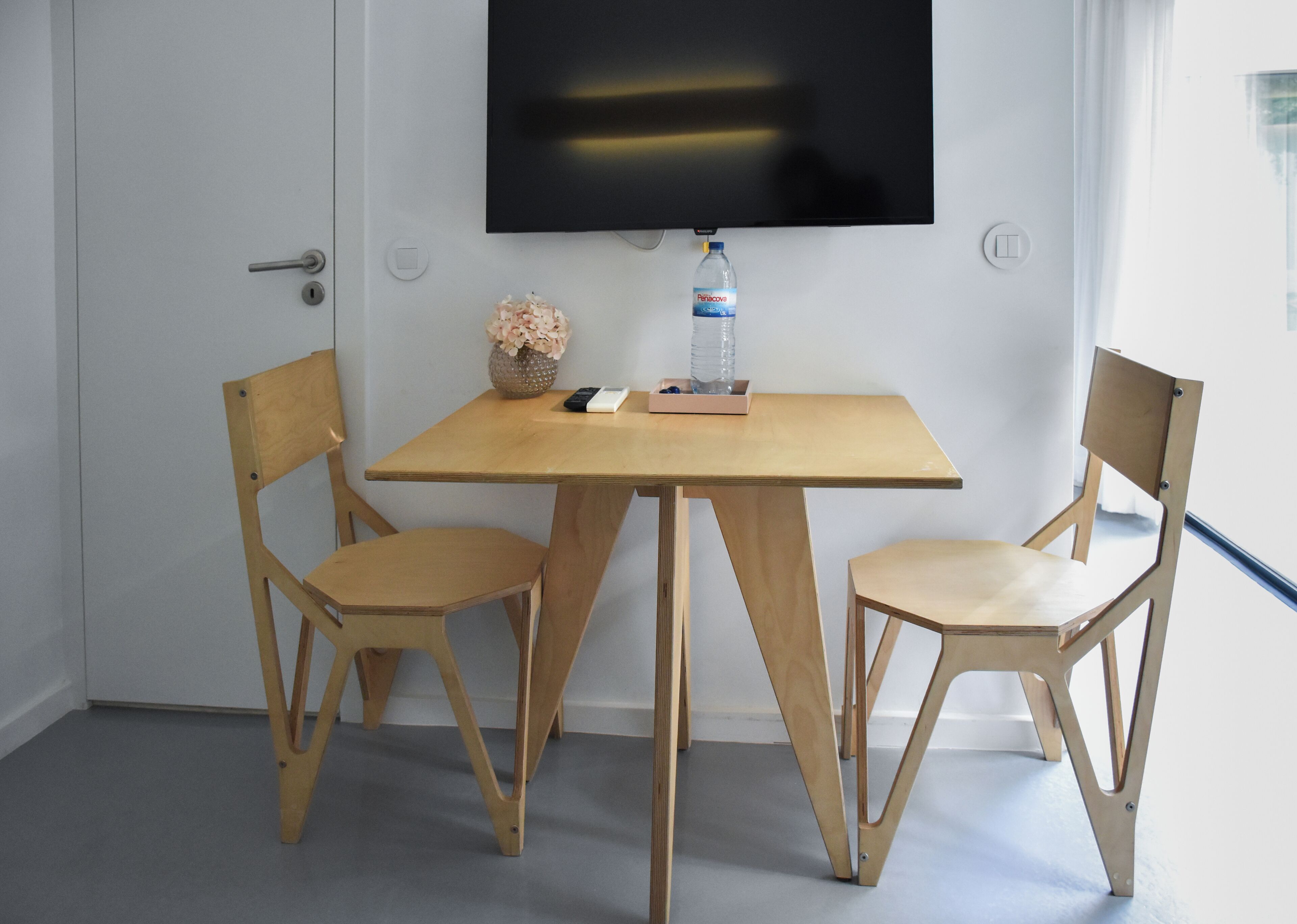 Ein Zimmer mit einem Holztisch, zwei Stühlen, einem an der Wand befestigten Fernseher, einer Wasserflasche und einer Vase mit Blumen.
