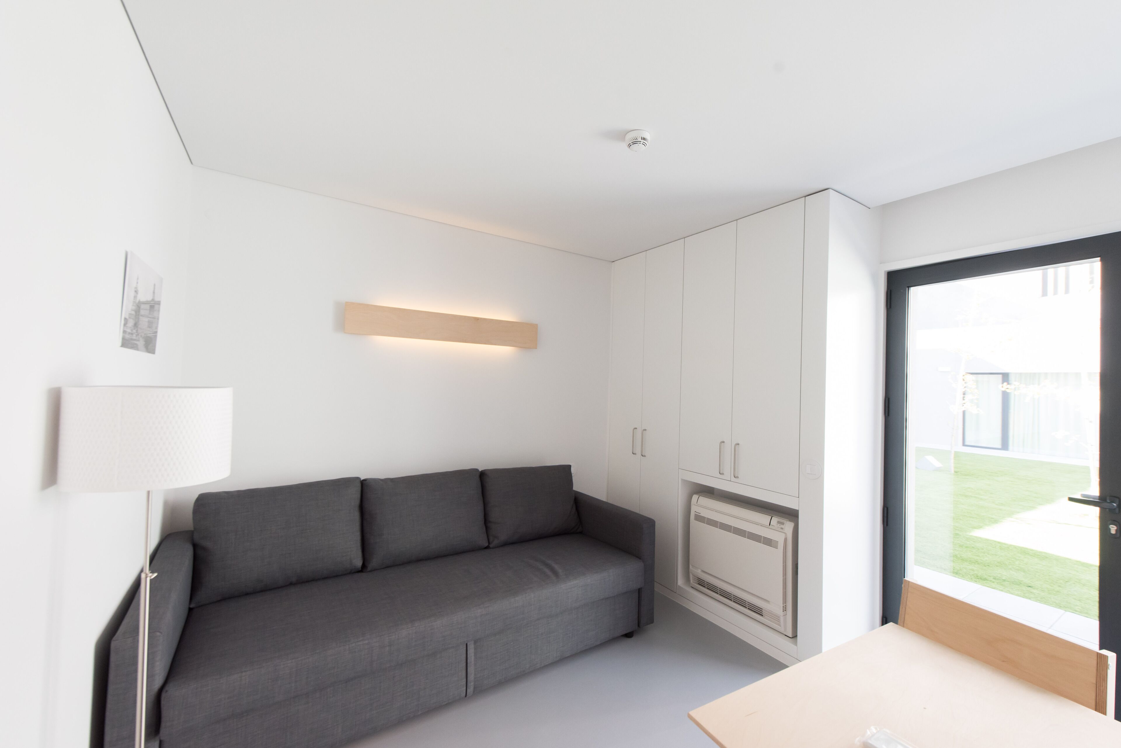 Egy modern, minimalista stílusú szoba, szürke kanapéval, egy kis asztallal és beépített fűtőtesttel.