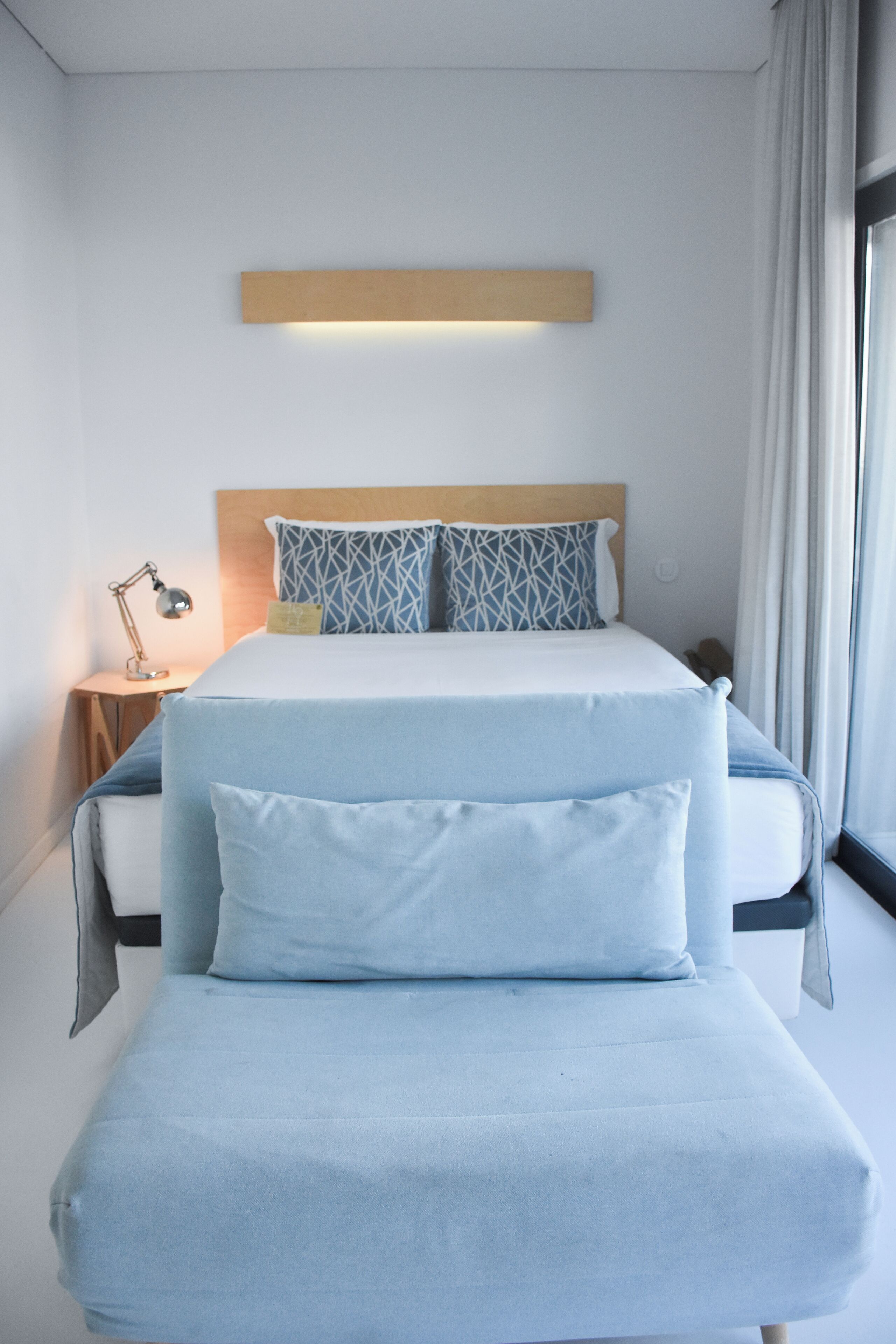 Habitación de hotel ordenada con cama, mesita de noche, lámpara y ventana con cortinas.