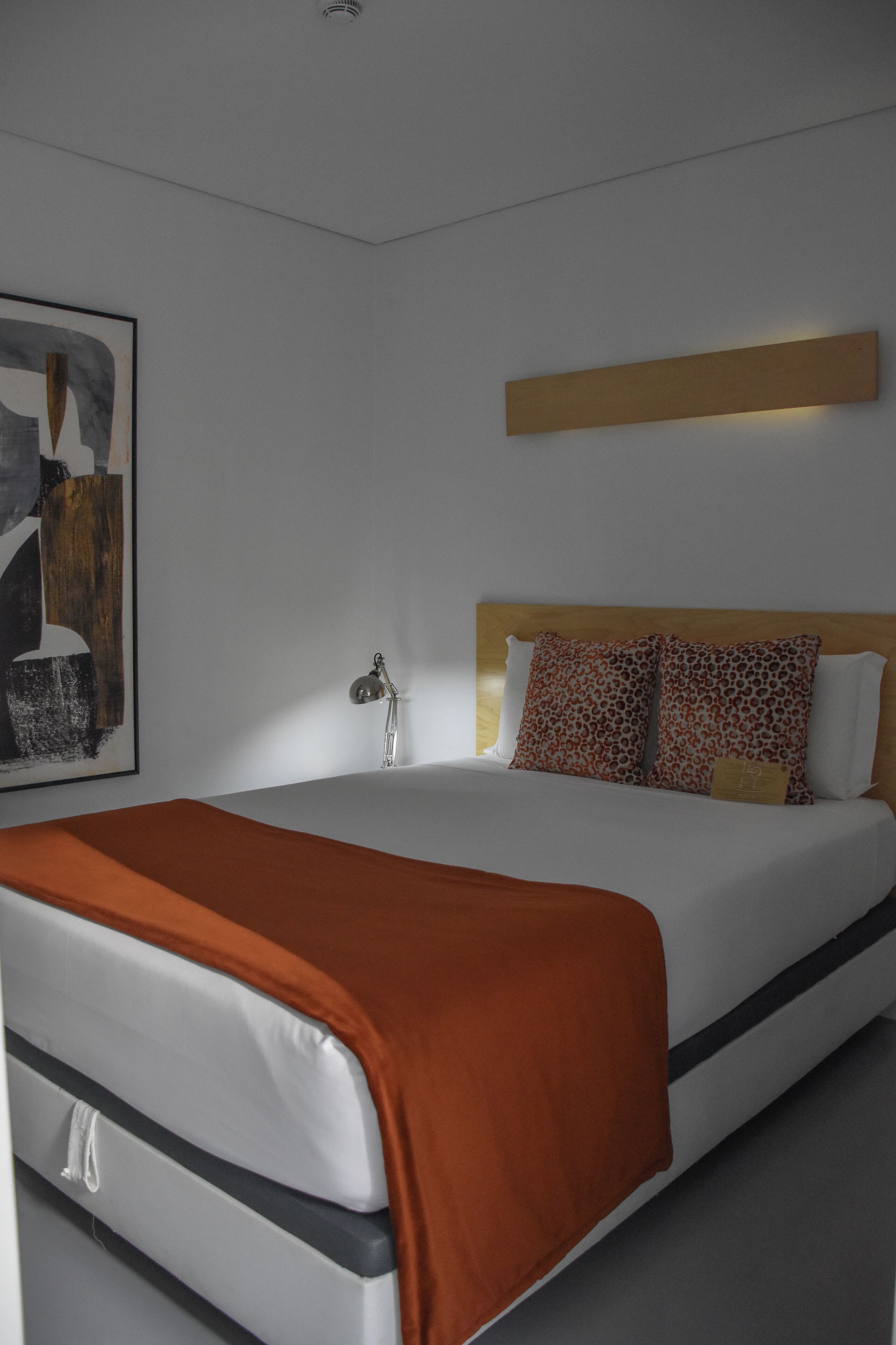 Una moderna camera d'hotel con un ampio letto, una testiera in legno, un comodino con una lampada e un quadro astratto incorniciato appeso al muro.
