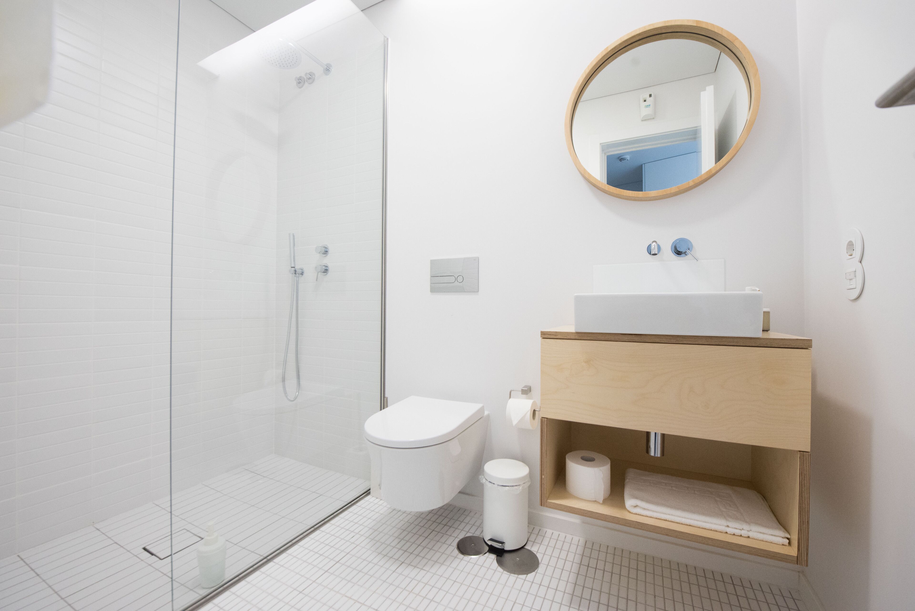 A modern bathroom with a glass shower enclosure, a white toilet, a wooden vanity with a sink, and a round mirror.